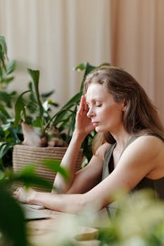 Adult woman sitting at home surrounded by plants, touching her temple with eyes closed, signifying a headache.